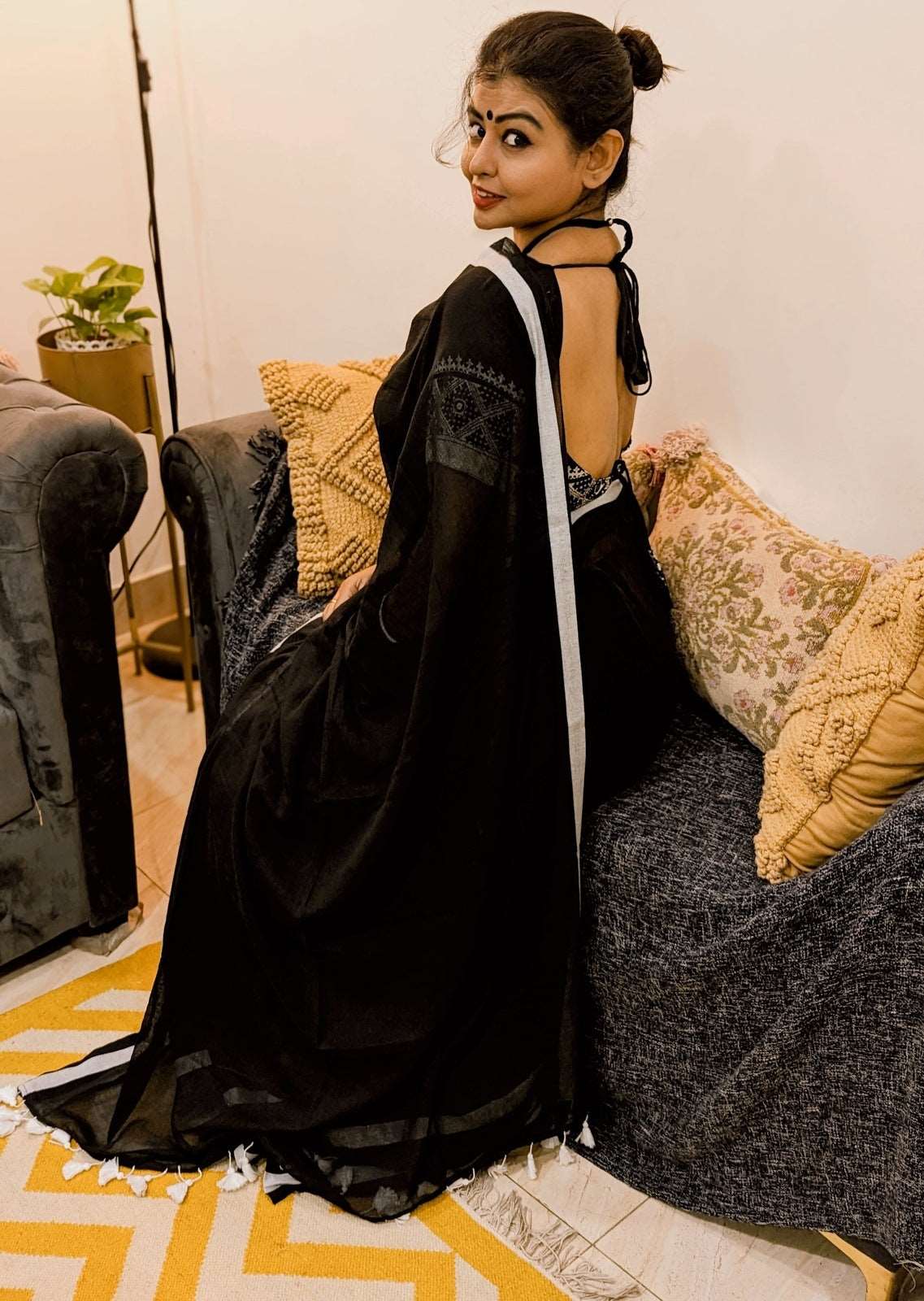 Black mul saree with delicate texture paired with a stylish black backless blouse worn by a model seated on a sofa.