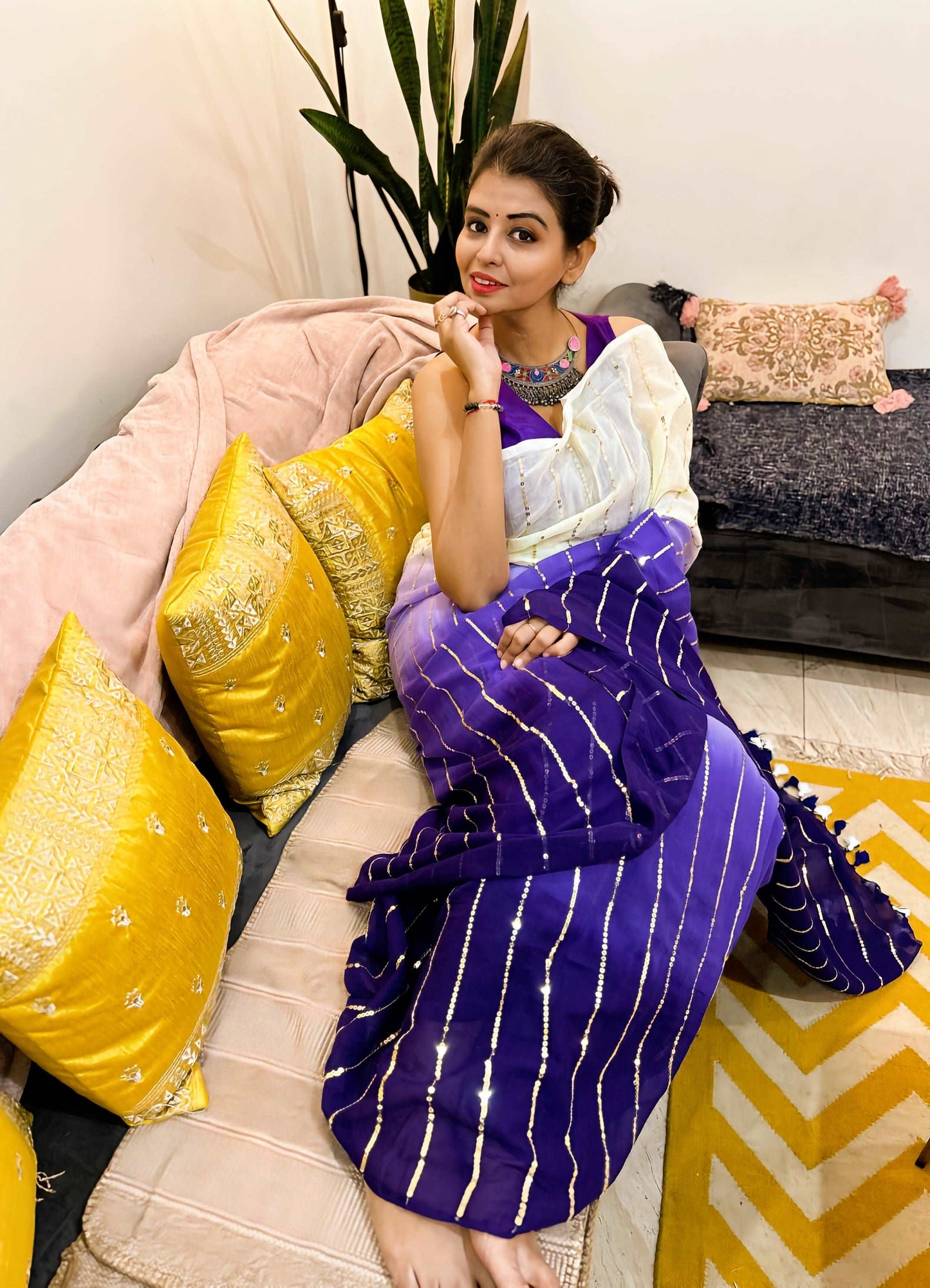 Woman wearing a purple Mul saree with white stripes and a white blouse, seated on a beige sofa with yellow cushions and a.