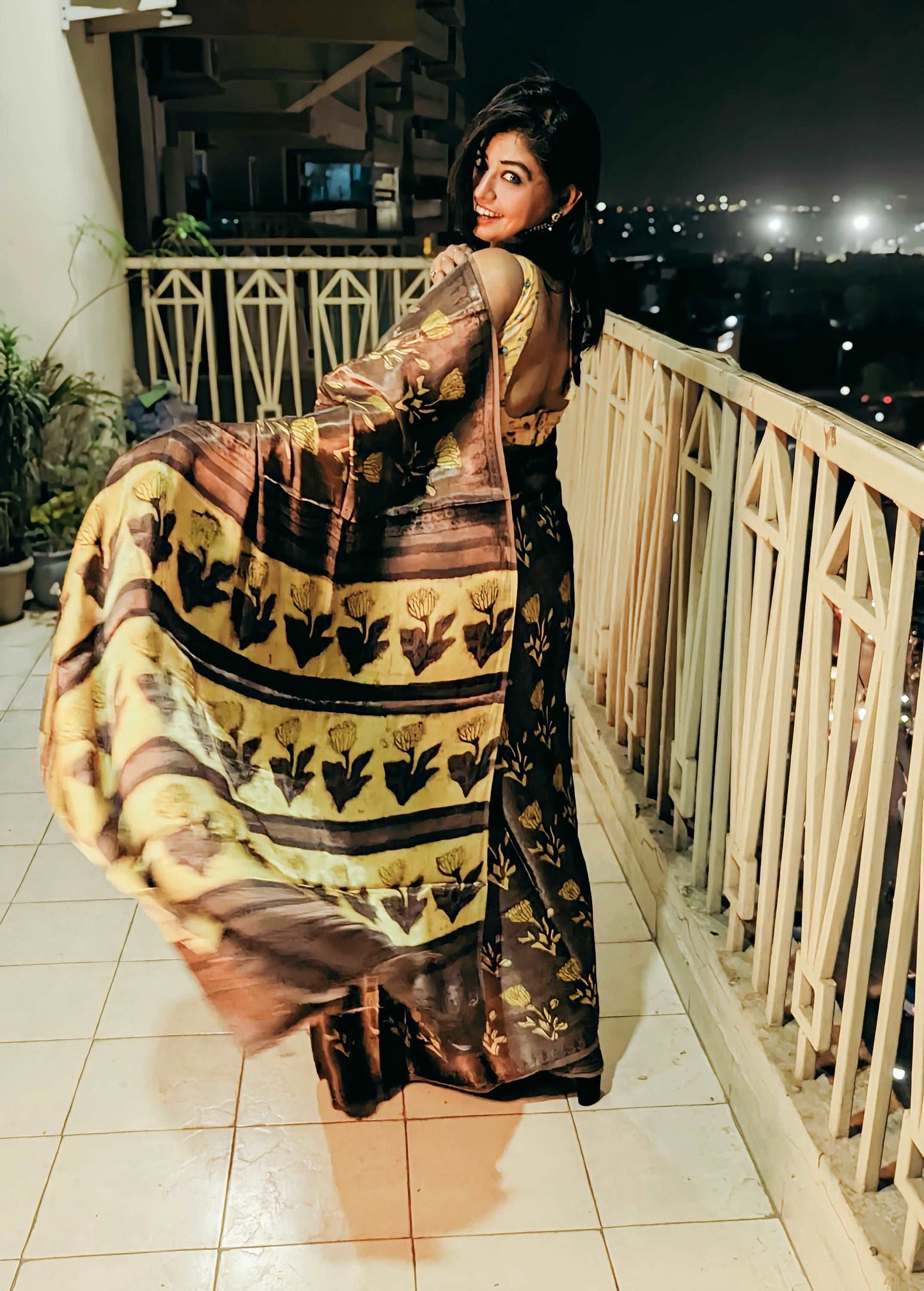 Yellow and brown Ajrakh handblock modal silk saree with floral and geometric patterns, worn by woman on balcony at night.