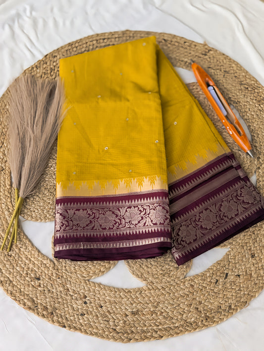 Mustard yellow silk saree with intricate maroon and silver Banarasi border detailing laid flat on a woven mat.