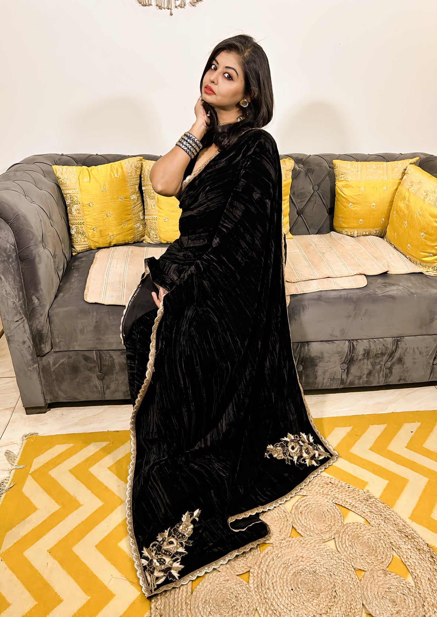 Black velvet saree with gold embroidered border worn by woman seated on gray sofa with yellow cushions.