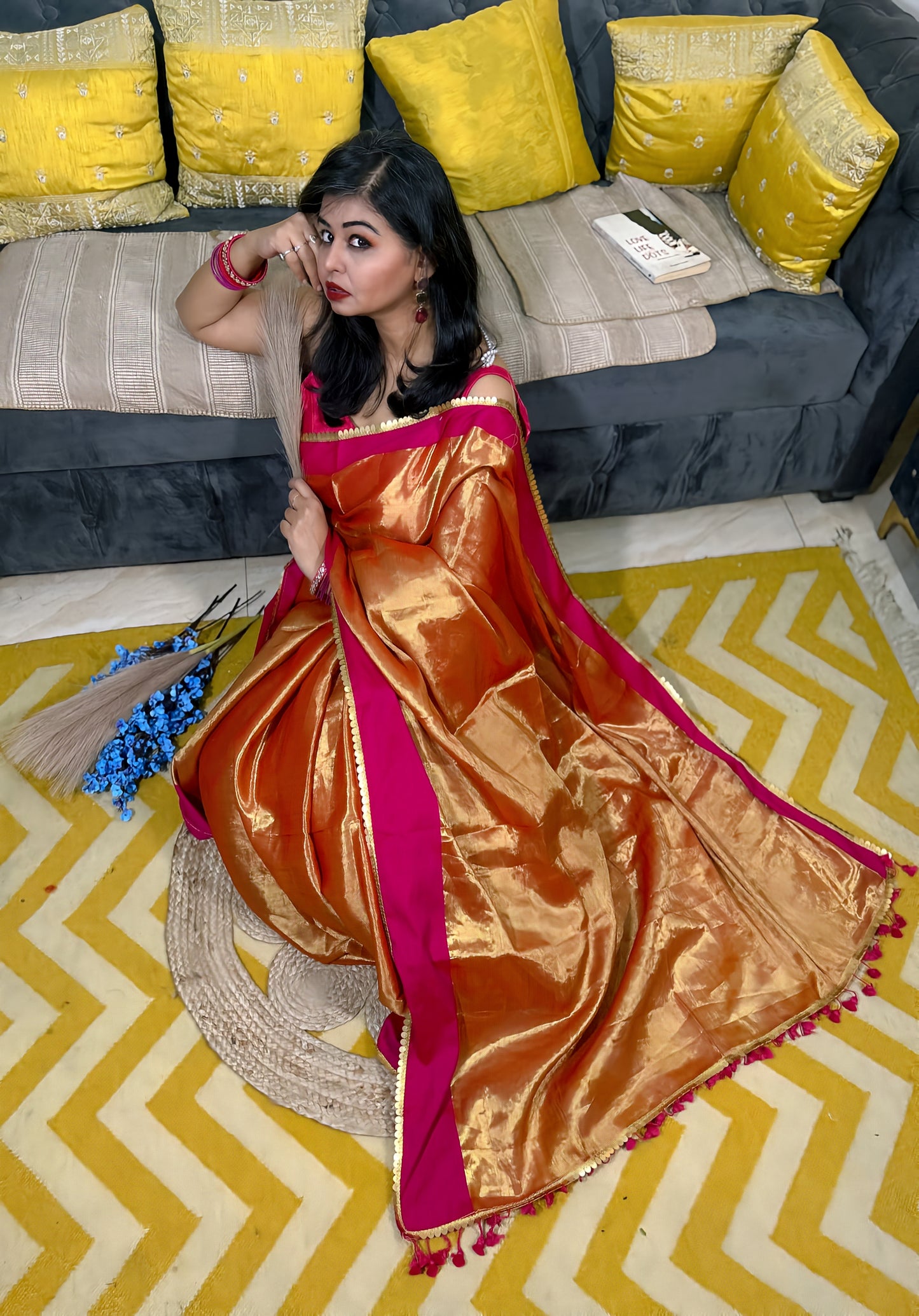 Rust-orange soft tissue saree with gold sheen and magenta border worn by woman sitting on yellow chevron rug.