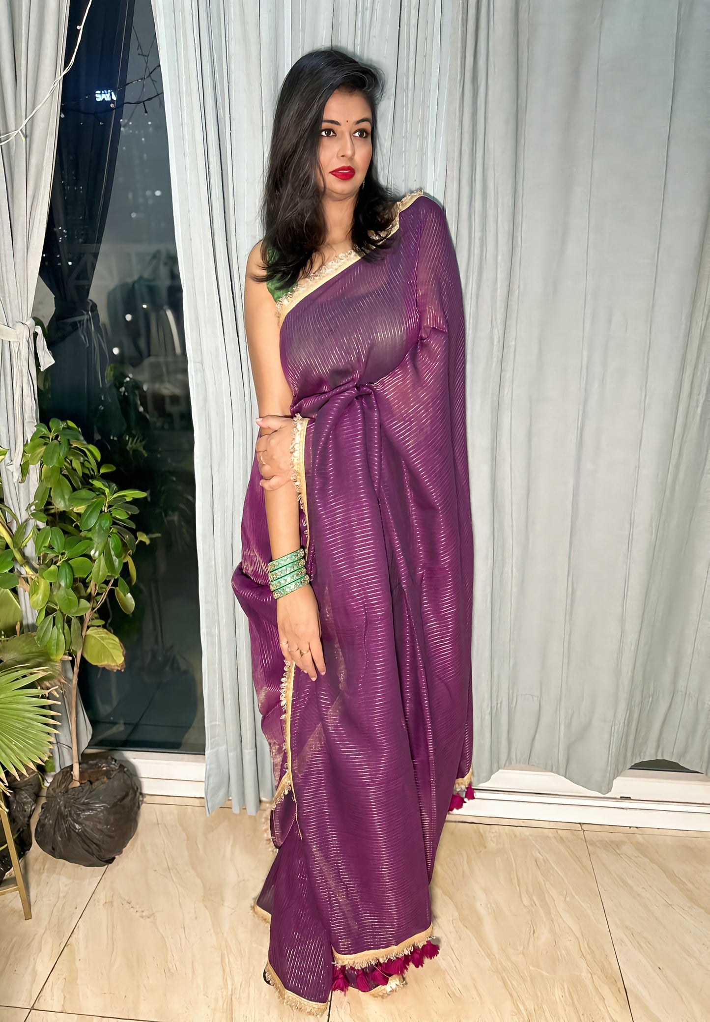 Deep purple mul zari saree with golden border and tassel detailing, styled with green bangles and red lipstick.