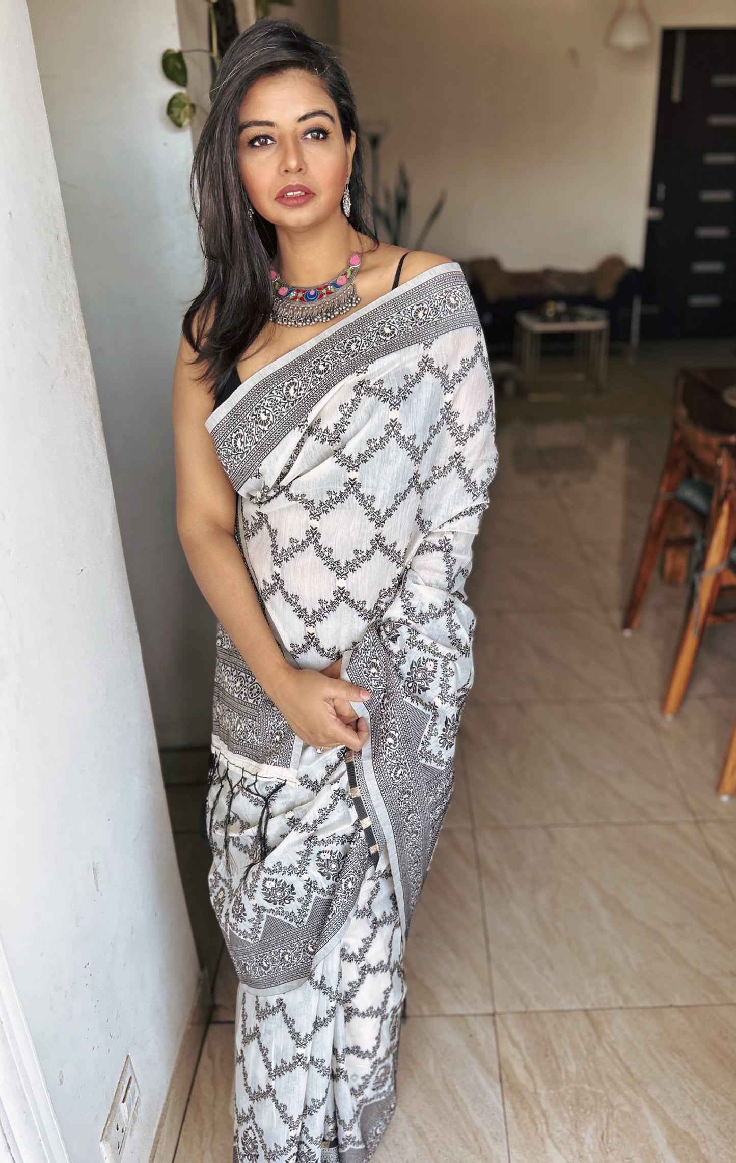 White and gray Bengal handloom saree with intricate floral motifs and geometric border worn with a blue necklace.