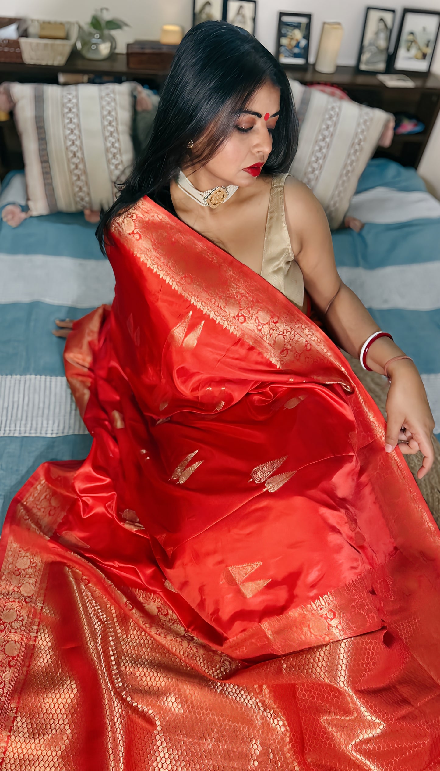 A woman showcasing a pinkish red Banarasi Gajji silk saree with golden zari buta and traditional karigari palla, sitting on a bed with a blue and white striped backdrop.