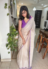 Off-white Bengal tissue saree with golden stripes and purple border draped on woman in sleeveless purple blouse.