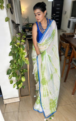 White mul cotton saree with green tie-dye patterns and blue sitara spark border worn with a blue sleeveless blouse.
