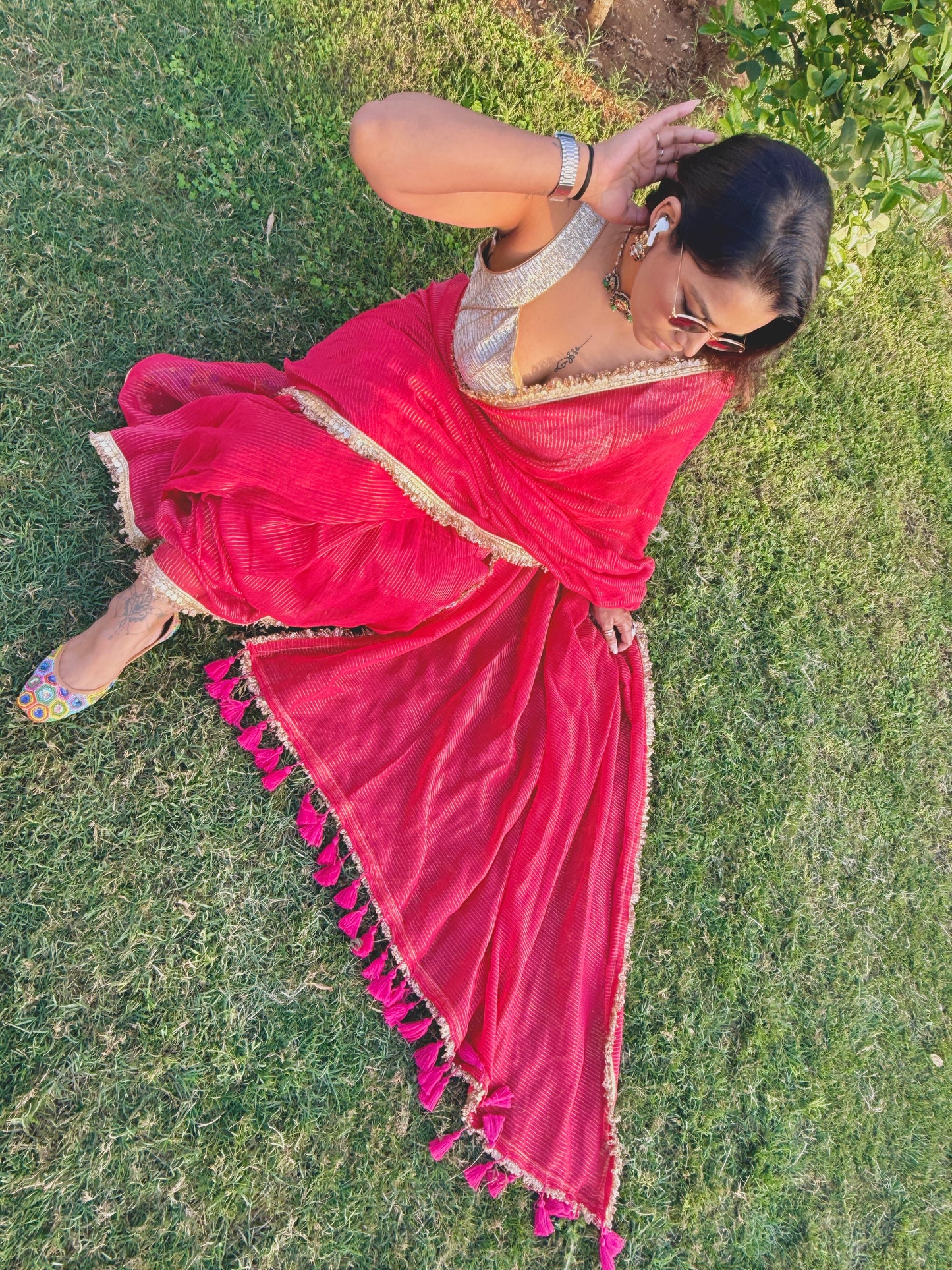 Red mul saree with tassel border paired with silver blouse worn outdoors on grass.
