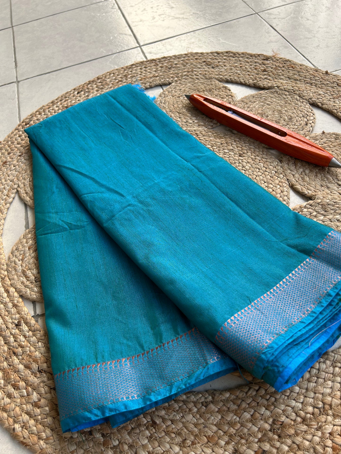 Teal blue cotton silk saree with silver woven border placed on round woven mat.