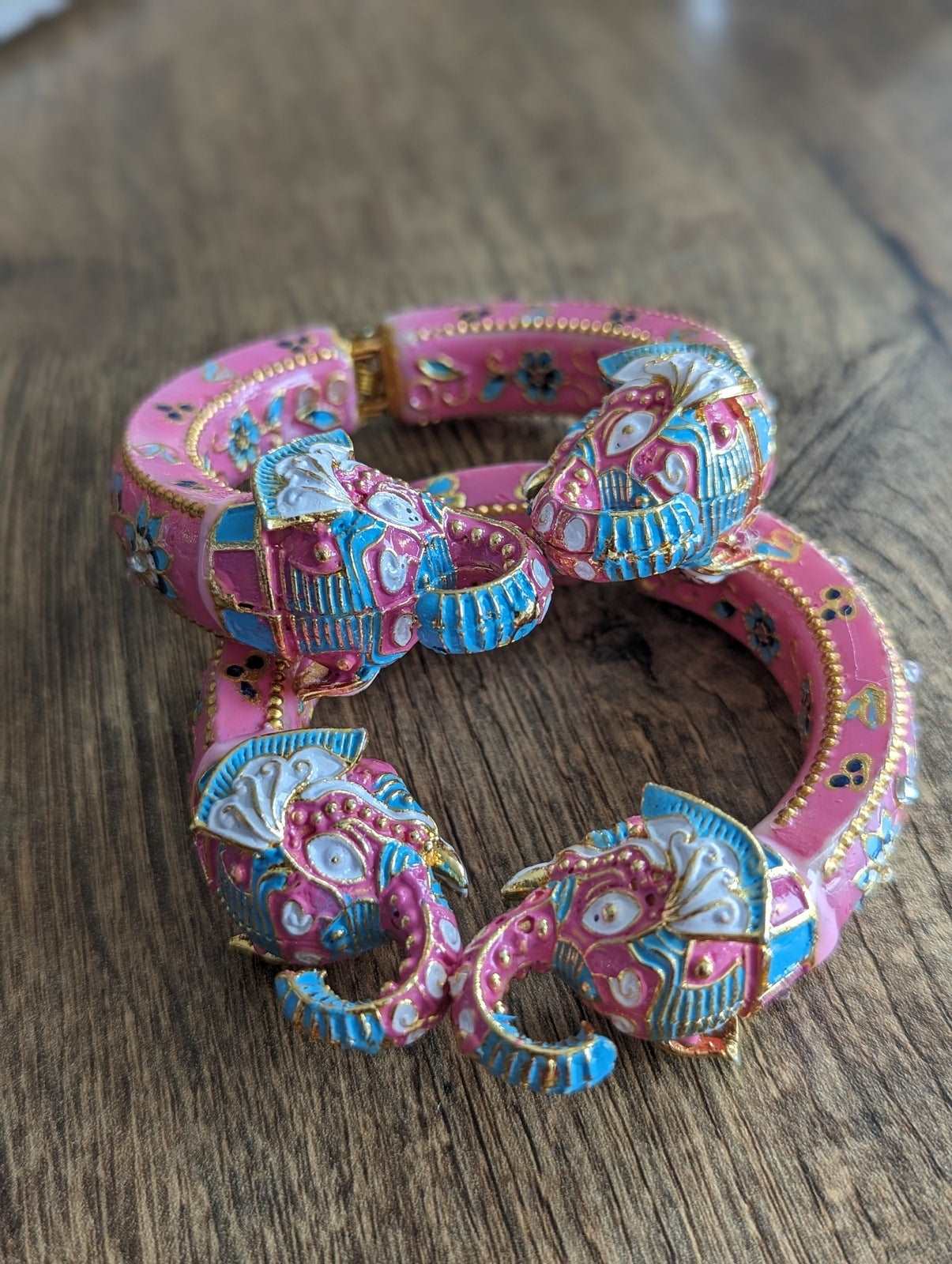 Pink and blue enamel bangles with intricate elephant head designs and gold accents on wooden background.