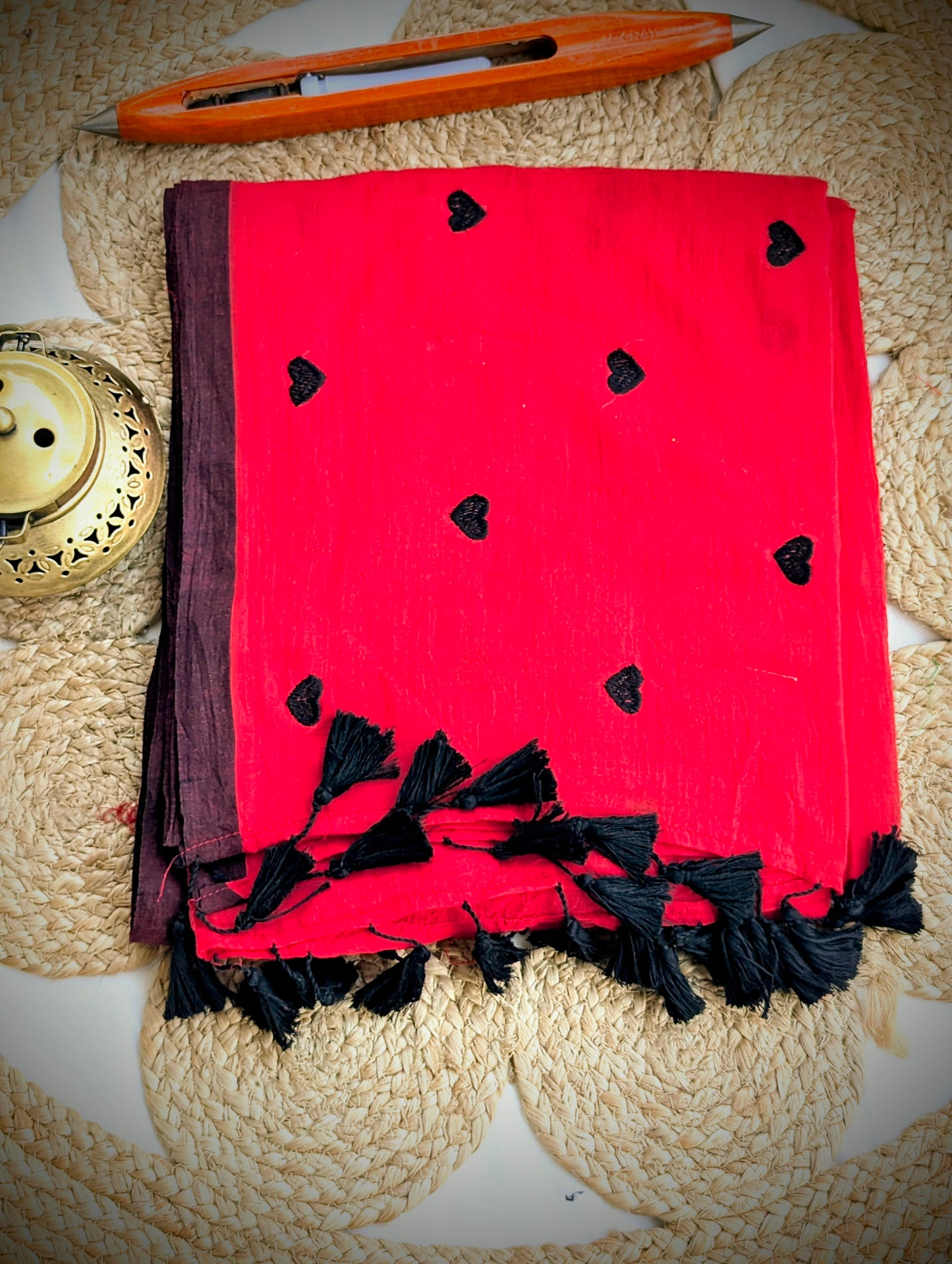 Crimson handloom Bengal cotton saree with black heart motifs and tassel fringe, folded on woven mat.