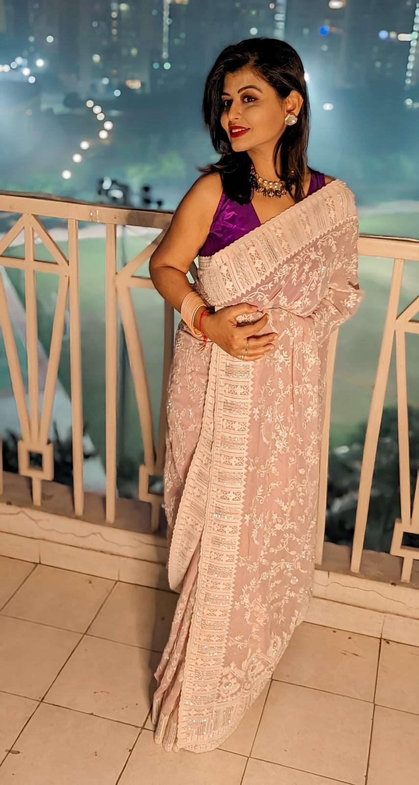 Pristine blush georgette saree with intricate thread embroidery worn with a purple blouse at night setting.