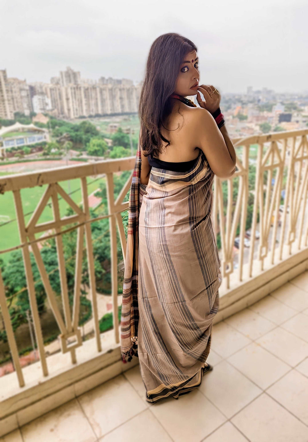 Beige and black Bhujodi Bengal handloom saree with subtle stripes, worn on a balcony overlooking a cityscape.