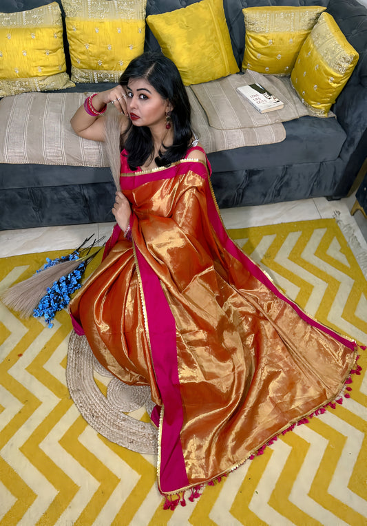 Rust-orange soft tissue saree with gold sheen and magenta border worn by woman sitting on yellow chevron rug.