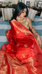 A woman showcasing a pinkish red Banarasi Gajji silk saree with golden zari buta and traditional karigari palla, sitting on a bed with a blue and white striped backdrop.
