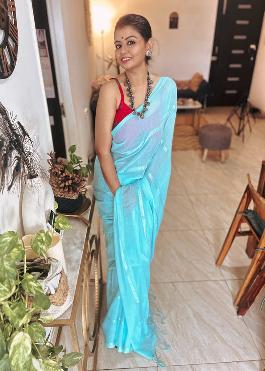Light blue silk saree with subtle sheen paired with red sleeveless blouse, worn by woman indoors.