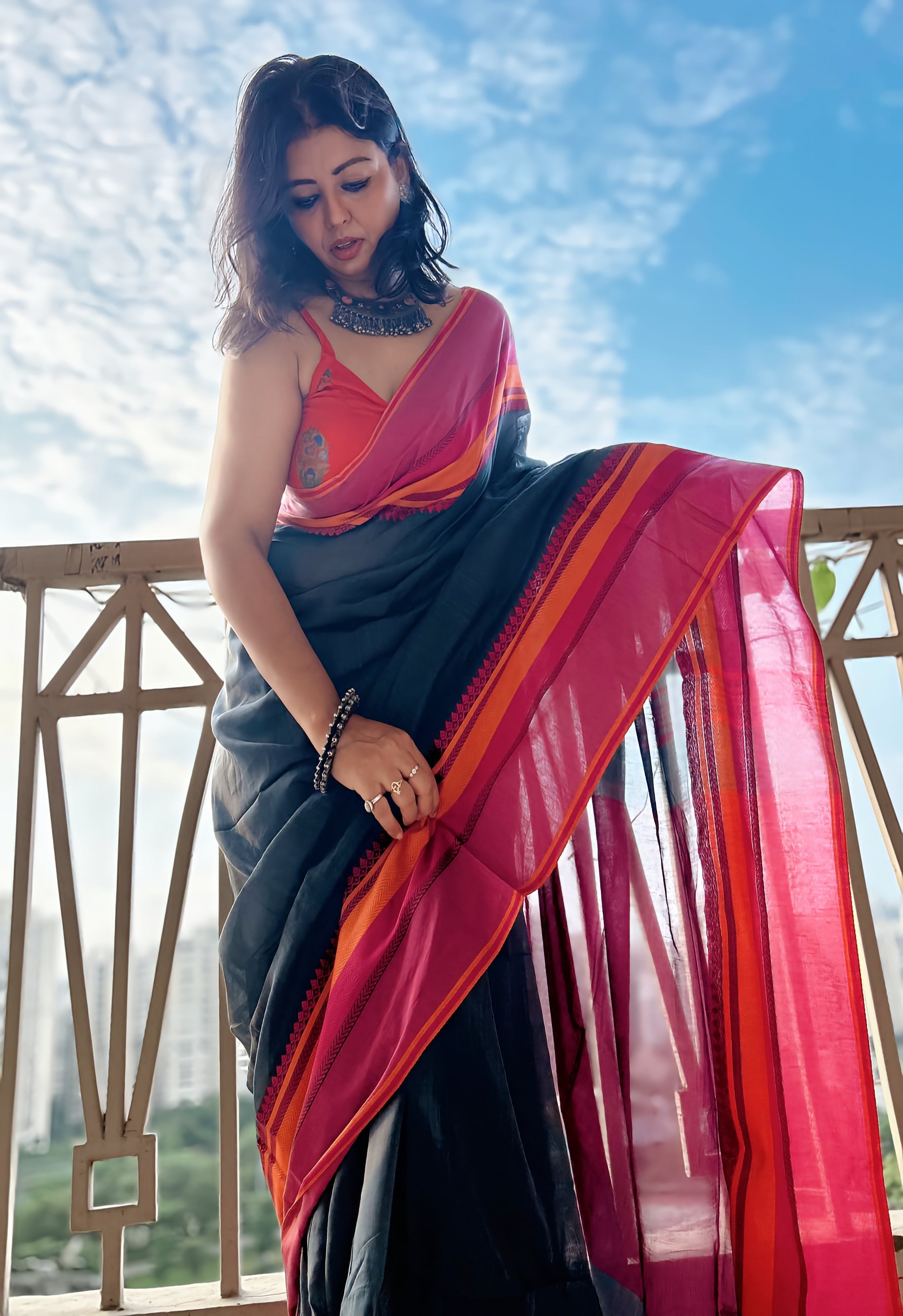 Black saree with bright pink and gold border, worn draped over shoulder, outdoors against blue sky background.