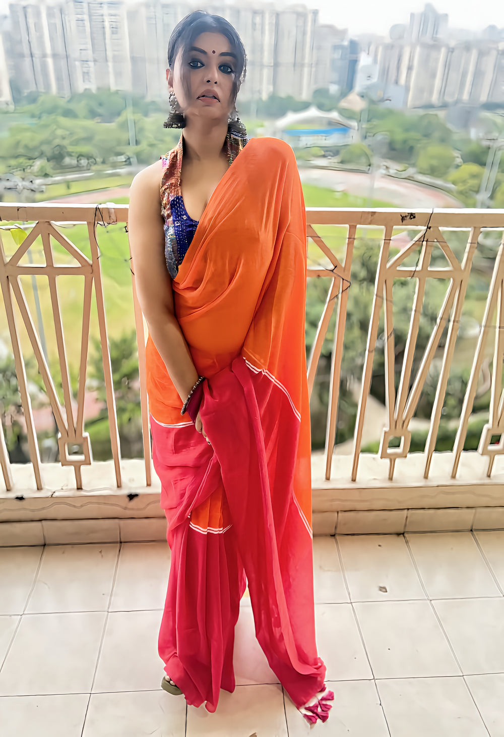 Bright orange and pink saree with halter-neck blouse worn by woman on balcony.