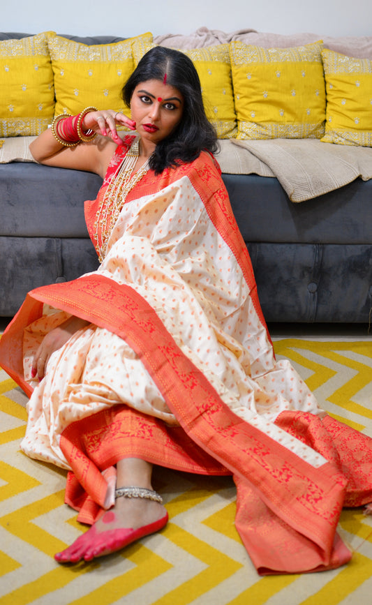 Off-white silk saree with orange floral motifs and broad orange border worn with matching blouse and pink jutti shoes.